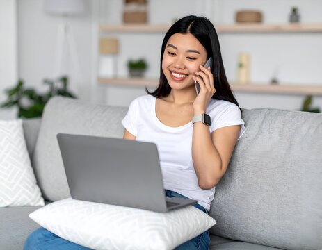 Smiling young Asian woman on sofa, comfortably managing calls and work on her laptop at home.