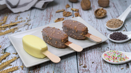 Delicious scene with three ice cream bars, including chocolate and vanilla flavors, served on a white plate with toppings, seeds, nuts, and colorful sprinkles. Rustic wooden background 