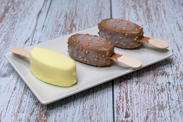 Delicious scene with three ice cream bars, including chocolate and vanilla flavors, served on a white plate with toppings, seeds, nuts, and colorful sprinkles. Rustic wooden background