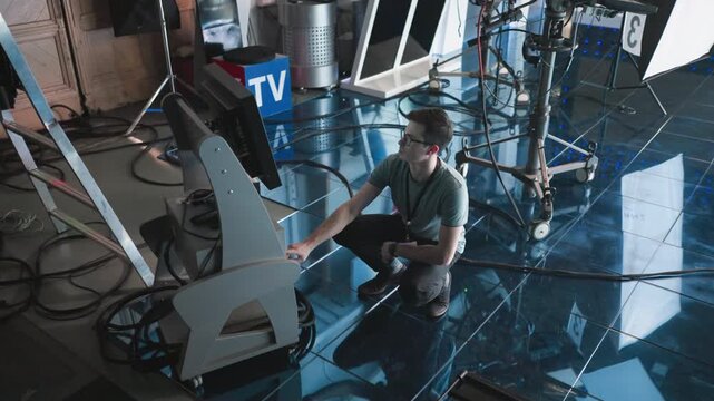 Man crouching and adjusting digital monitor and timer settings in modern studio, professional production environment with filming equipment, focused on workspace, tech action in progress