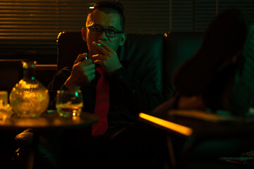 Man sits on a sofa in the room, lighting a cigarette, holding a glass of whiskey, drinking with a frustrated expression on his face.
