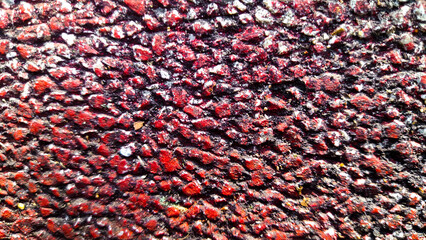 A close-up of an abstract, textured surface of red asphalt, showcasing its rough and uneven pattern with dark crevices.