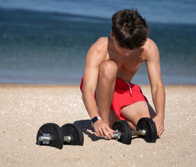 Shirtless athlete assembling dumbbells on a sandy beach, getting ready for an outdoor workout by...