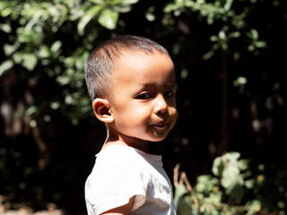 Side portrait of a handsome child, adorable boy in white t-shirt looking through his shoulder at camera, with summer natur background. Copy ad space.