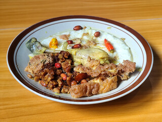 A dish of chicken porridge with yellow sauce, chicken pieces, omelet, orek tempeh and fried peanuts, served on a brown enamel plate. Chicken porridge, traditional Indonesian food.