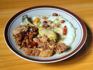 A dish of chicken porridge with yellow sauce, chicken pieces, omelet, orek tempeh and fried peanuts, served on a brown enamel plate. Chicken porridge, traditional Indonesian food.