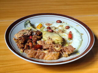 A dish of chicken porridge with yellow sauce, chicken pieces, omelet, orek tempeh and fried peanuts, served on a brown enamel plate. Chicken porridge, traditional Indonesian food.