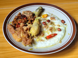 A dish of chicken porridge with yellow sauce, chicken pieces, omelet, orek tempeh and fried peanuts, served on a brown enamel plate. Chicken porridge, traditional Indonesian food.