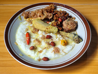 A dish of chicken porridge with yellow sauce, chicken pieces, omelet, orek tempeh and fried peanuts, served on a brown enamel plate. Chicken porridge, traditional Indonesian food.