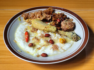A dish of chicken porridge with yellow sauce, chicken pieces, omelet, orek tempeh and fried peanuts, served on a brown enamel plate. Chicken porridge, traditional Indonesian food.