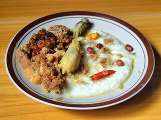 A dish of chicken porridge with yellow sauce, chicken pieces, omelet, orek tempeh and fried peanuts, served on a brown enamel plate. Chicken porridge, traditional Indonesian food.