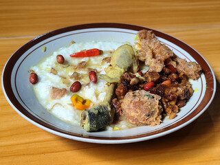 A dish of chicken porridge with yellow sauce, chicken pieces, omelet, orek tempeh and fried peanuts, served on a brown enamel plate. Chicken porridge, traditional Indonesian food.