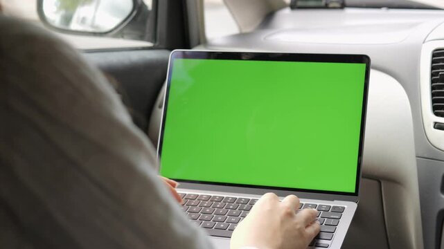 Person is using a laptop in a car. The laptop screen is green. The person is typing on the keyboard