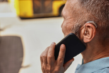 Senior man with hearing aid speaking phone in urban street, side view, close-up of ear with BTE...