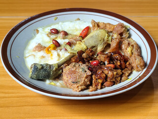 A dish of chicken porridge with yellow sauce, chicken pieces, omelet, orek tempeh and fried peanuts, served on a brown enamel plate. Chicken porridge, traditional Indonesian food.