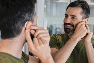 Naklejka na ściany i meble Man with hearing problems tests out his new behind-the-ear hearing aid, smiling as he observes himself in home mirror. Audiology