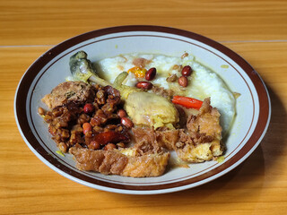 A dish of chicken porridge with yellow sauce, chicken pieces, omelet, orek tempeh and fried peanuts, served on a brown enamel plate. Chicken porridge, traditional Indonesian food.