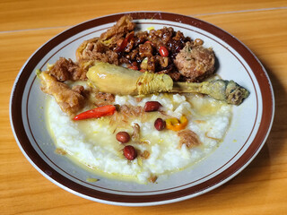 A dish of chicken porridge with yellow sauce, chicken pieces, omelet, orek tempeh and fried peanuts, served on a brown enamel plate. Chicken porridge, traditional Indonesian food.