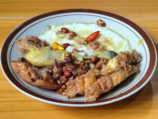 A dish of chicken porridge with yellow sauce, chicken pieces, omelet, orek tempeh and fried peanuts, served on a brown enamel plate. Chicken porridge, traditional Indonesian food.