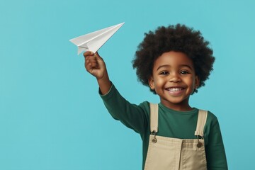 Young African American boy with curly hair joyfully holding a paper airplane against a vibrant blue background, showcasing creativity and childhood imagination in a playful atmosphere