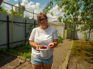 Smiling woman holding white bucket full of freshly picked cherries showing few berries in hand...
