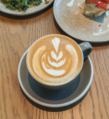 A cup of cappuccino with a decorative latte art design is served on a saucer on a wood table.