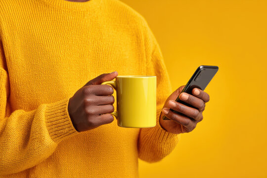 Close-up of person wearing yellow sweater, holding ceramic coffee mug in one hand and smartphone in other against solid yellow background - Powered by Adobe