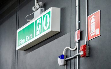 Green fire exit sign, emergency light, fire extinguisher symbol sign and red alarm siren with flashing strobe light mounted on the wall. Fire alarm system and evacuation guiding equipment in building.