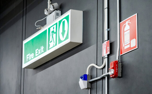 Green fire exit sign, emergency light, fire extinguisher symbol sign and red alarm siren with flashing strobe light mounted on the wall. Fire alarm system and evacuation guiding equipment in building.