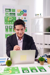 Asian adult businessman analyzing ESG data on laptop in modern office promoting corporate...