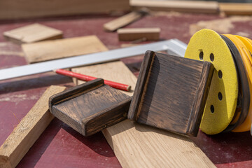 Woodworking tools and materials laid out on a workbench, including a yellow orbital sander, red carpenter's pencil, various wood pieces, and a metal square ruler.