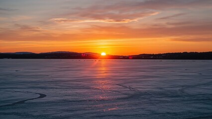 Fototapeta premium Golden hues illuminate a frozen lake at sunset, highlighting the striking contrast between the warm sky and chilly winter landscape.