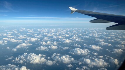 Fototapeta premium Quiet flight window outlook showing plane wing, billowy clouds beneath, and clear azure sky above, stretching over a vast patterned scenery.
