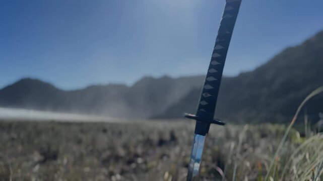 Long Japanese samurai sword katana is positioned in a windy grass field ground. Strong sunlight.