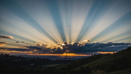 Blue sky adorned with floating clouds. Landscape background. Evening sun descending. Warm and tranquil evening. Sun setting beyond slopes and valleys. Sunshine piercing through cloud cover.