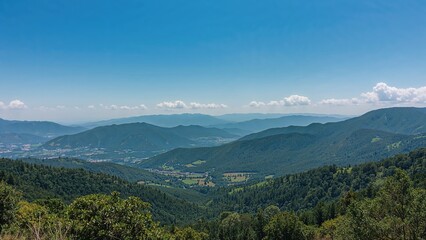 Fototapeta premium Broad perspective of rich, green mountain ranges and valleys beneath a sunny sky, ideal for promoting outdoor journeys and nature appreciation.