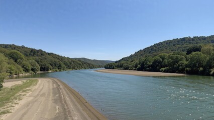 Calm river waters beside gently inclined sandy banks surrounded by rich plant life, with forested hills in the background beneath a pristine blue sky
