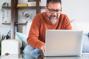 Middle-aged man working from home on laptop in living room, sitting on sofa with table, focused and stressed freelancer dealing with bills, household economy, deadlines and responsibilities
