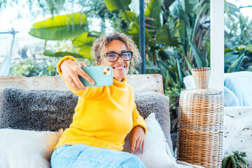 Middle-aged woman using smartphone in garden at home, wearing yellow oversized sweater, smiling and...