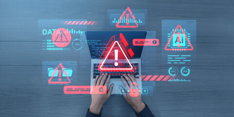 Cybersecurity alert concept showing hands on a laptop keyboard with warning graphics and data breach icons on digital interface overlays on wooden desk.