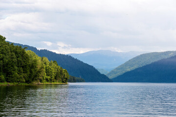 A tranquil mountain lake reflects the surrounding lush green hills and distant peaks under a cloudy sky