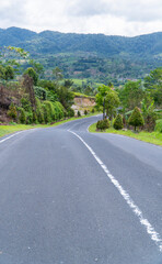 Winding asphalt road through lush green hills with mountain backdrop under cloudy sky