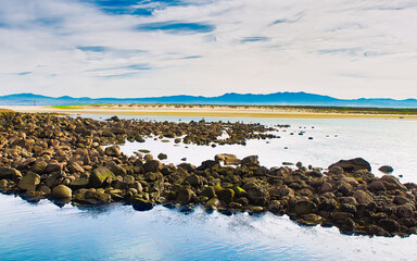 Mossy rocks in the foreground frame a serene beach and mountain view on the C&iacute;es Islands, Galicia, Spain. A perfect blend of texture, depth, and natural beauty.