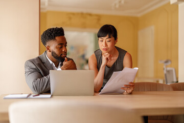 Focused professionals discussing project details and reviewing documents with laptop on desk in...