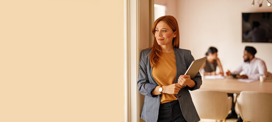 Thoughtful businesswoman with laptop looking away confidently while leaning on wall with colleagues in the background