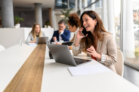 Cheerful businesswoman talking over smart phone while sitting with laptop at conference table during business meeting - Powered by Adobe