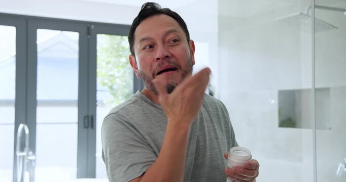 Starting daily grooming mature adult Asian man twisting off lid at bathroom mirror applying cream