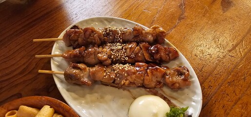 Grilled chicken skewers glazed with sweet soy sauce and sprinkled with sesame seeds, served with a soft-boiled egg on a ceramic plate. Traditional Korean izakaya-style dish on a rustic wooden table.