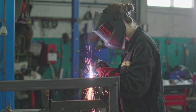 Skilled female mechanic in a protective helmet welding a metal frame in an auto repair garage