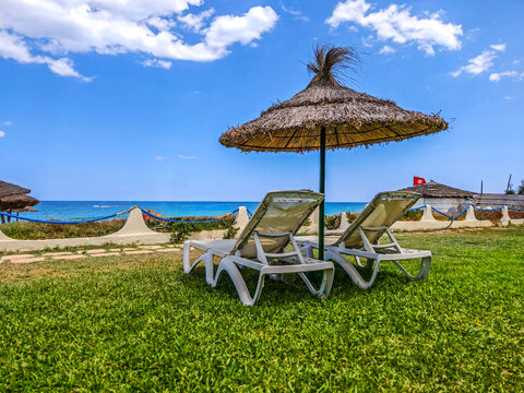 Sunloungers sit beneath a rustic straw umbrella on a vibrant green lawn overlooking the sparkling blue ocean under a clear sky. sousse, tunisia